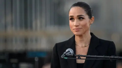 Getty Images Meghan Markle stands behind a microphone wearing a simple charcoal jacket and top, a gold necklace hangs across her chest. Her dark hair is pulled back in a bun.