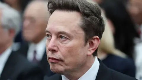 Getty Images Elon Musk looks sideways as he wears a dark suit, white shirt and grey tie, with other people visible in the background.