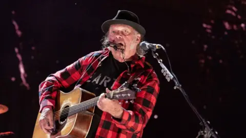 Getty Images Neil young wears a red and black checked shirt and plays a guitar.