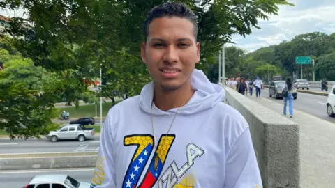 A man wearing a white hoodie and chain. The letter V in Venezuela's national colours and a map of the country is in the front of his hoodie. Behind him is a public street with cars and walkers in view.
