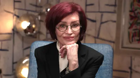 BBC Sharon Osbourne poses with her chin resting on her hand, against a colourful background