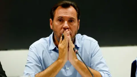 EPA A middle-aged man in a light blue button-down shirt, placing his hands together in a prayer position in front of his mouth while sitting in front of a microphone at a press conference.