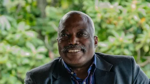 Getty A middle aged man with a greying moustache smiles into the camera wearing a blue coat and a blue and red striped shirt sitting in front of a blurry nature background