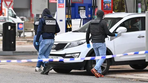 Getty Images Forensic officers examine evidence at the site of a petrol station where a gunman opened fire from a vehicle, killing one person and injuring four others, in Trappes.
