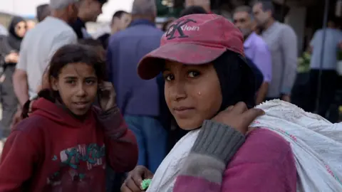 Neha Sharma/BBC A girl wearing a pink baseball cap with a white sack over her shoulder looks at the camera, with a second girl behind her and a crowd of people behind them standing further away