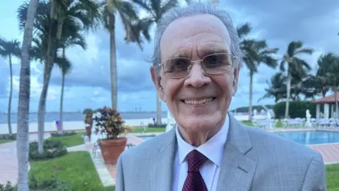BBC Juan Omar Sixto stands in front of a swimming pool and Biscayne Bay in Florida. He is smartly dressed in a suit and tie