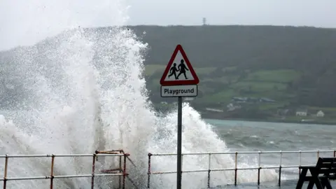 Reuters A wave crashing up onto a path with a metal railing and a black chair. There is a red and white triangular playground sign.