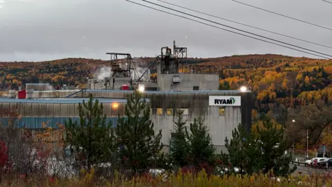 BBC/Jessica Murphy An image showing a part of the RYAM plant in Temsicaming, surrounded by red and orange fall trees. The building is grey with a sign in right corner that features the RYAM logo. Smoke is billowing out the ceiling. 