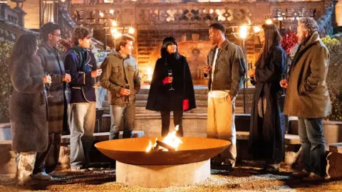 BBC/Studio Lambert/Euan Cherry The seven Celebrity Traitors finalists standing around a fire pit outside the castle listening to Claudia Winkleman