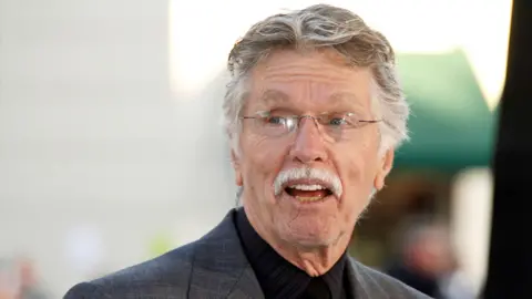 REUTERS/Fred Prouser/File Photo Actor Tom Skerritt is seen with his mouth open, apparently talking, and smiling. He is wearing a black buttoned-up shirt, rimless glasses and a grey suit coat. He appears to be outside. 
