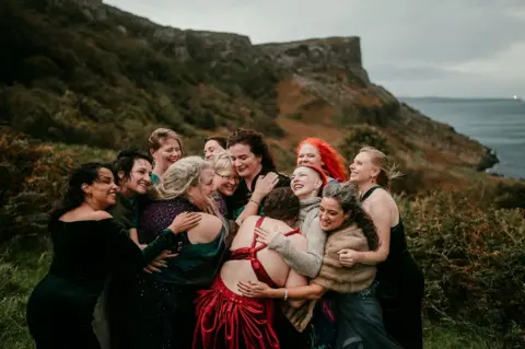 Rob Dight / Epic Love Photography A group of 10 women stand in a close embrace on a grassy hillside overlooking the sea. Several wear formal dresses in dark and red colours. Behind the group, rugged coastal cliffs rise under an overcast sky.