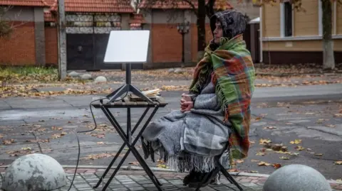 Reuters A man, with at least three blankets around him and a drink in his hand, sits on a chair on the side of a street. Next to him is a small picnic table with a starlink terminal on it.